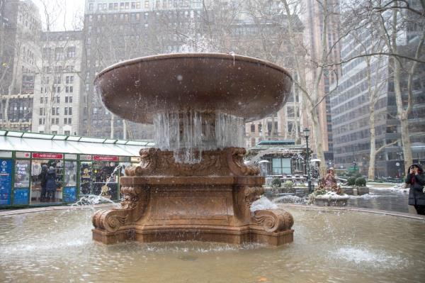 An iced-over Bryant Park fountain seen with popup shops in the background, as flurries fall and visitors snap pics with hoods up.